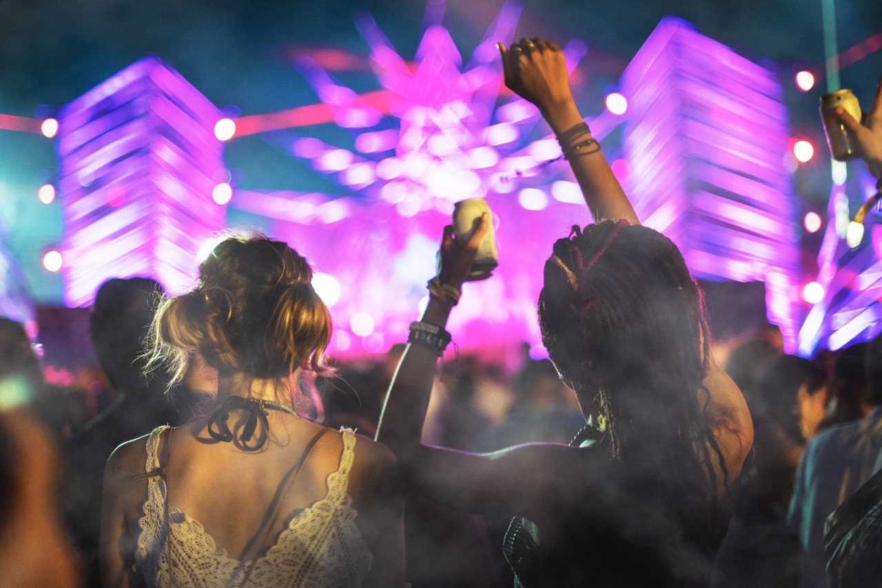 Pessoas curtindo a música em um festival ao ar livre, com luzes neon vibrantes e fumaça no ar criando uma atmosfera energética.