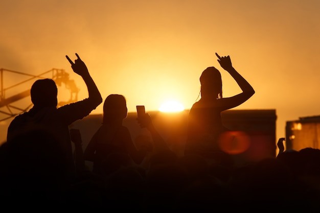 Silhuetas de três pessoas em meio a uma multidão curtem um show ao ar livre durante o pôr do sol. Duas delas levantam as mãos fazendo o gesto clássico do rock, enquanto outra segura um celular. O céu alaranjado e a iluminação natural criam uma atmosfera vibrante, representando a paixão pela música ao vivo e a energia dos festivais.