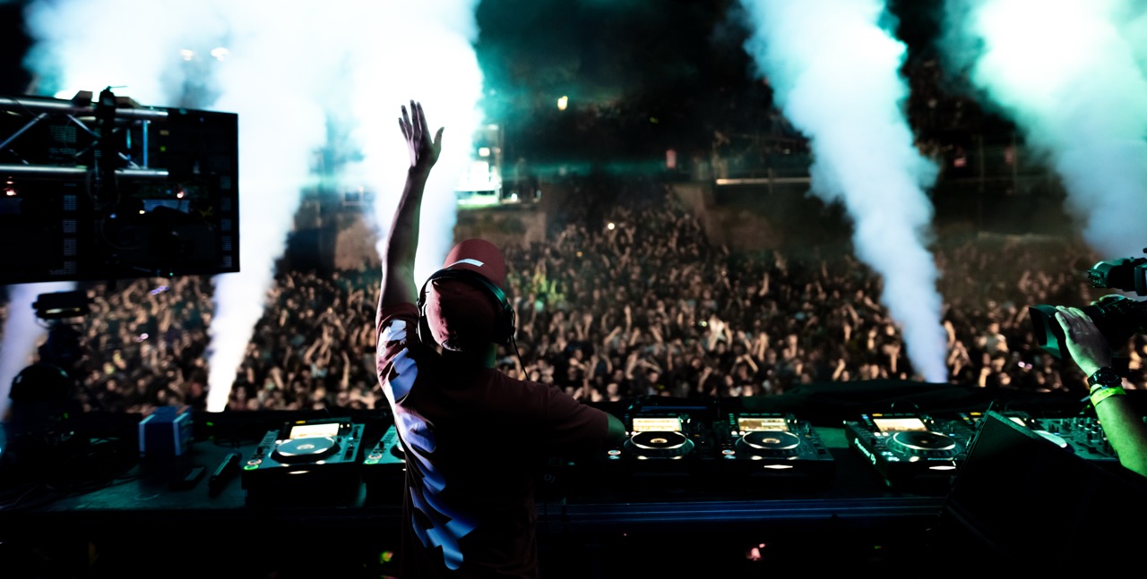 DJ com fones de ouvido tocando música ao vivo para uma multidão animada em um grande festival noturno, com efeitos de fumaça e luzes no palco.