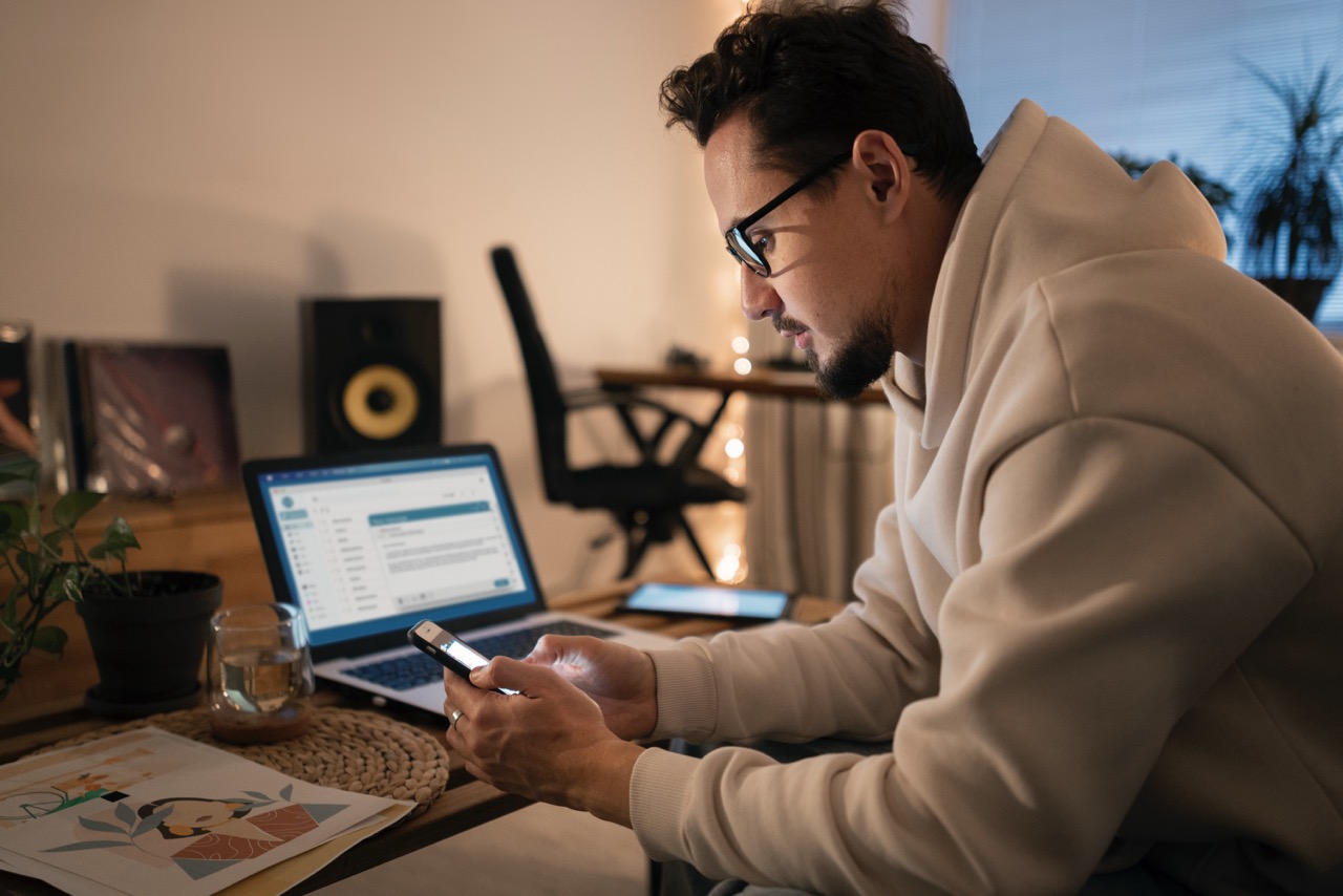 Homem usando notebook e celular em ambiente doméstico à noite, trabalhando estratégias de branding pessoal e gerenciamento de marca digital.