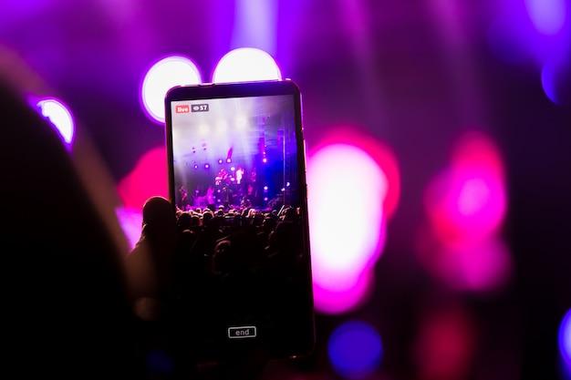 Pessoa filmando um show com o celular em modo live, representando o impacto das redes sociais nas transmissões ao vivo e no compartilhamento instantâneo de experiências musicais.
